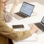 Two women collaborate on financial data analysis with laptops in an office setting.